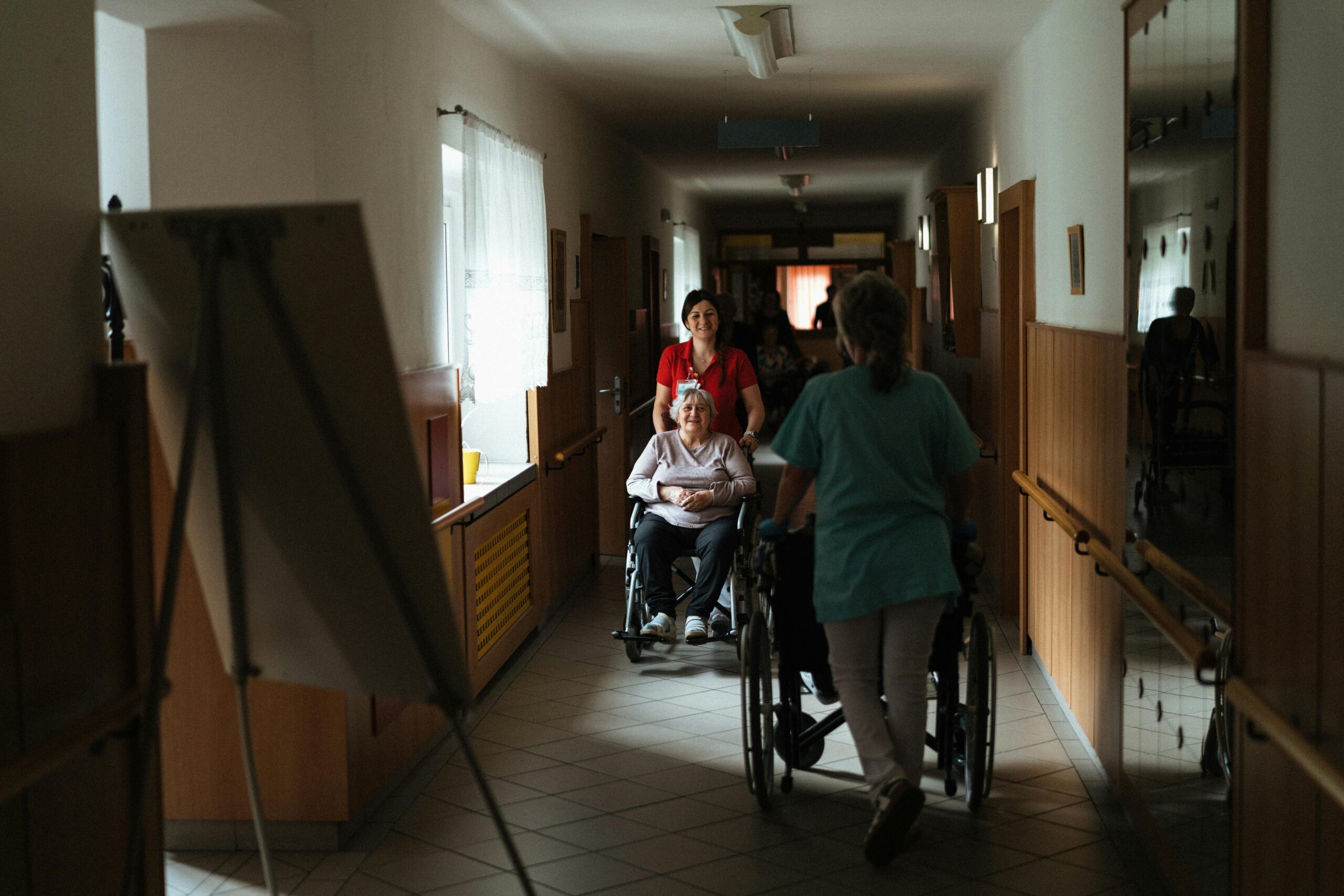 Caregiver assisting elderly woman in a wheelchair through a retirement home corridor.